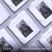 Футболка єнот пілот, біла, на моделі, з фронтального ракурсу, фон світло-фіолетовий, принт з єнотом у пілотському шоломі з сигаретою, на фоні літаки та вибухи.