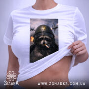 Футболка єнот пілот, біла, на плоскій поверхні, з верхнього ракурсу, фон світлий, принт з єнотом у пілотському шоломі з сигаретою, на фоні літаки та вибухи, з етикеткою.