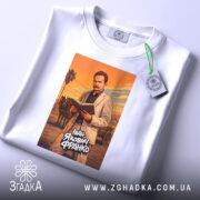 Футболка Іван Франко білого кольору, розкладена на поверхні. Принт зображає чоловіка в світлому костюмі, який читає книгу, на фоні оранжевого заходу сонця. На футболці видно етикетку з зеленим шнурком, що додає акценту. Фон нейтральний, світло-сірий.