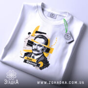 Футболка Іван Франко, біла, на столі, складена, з принтом портрета Івана Франка, фон світло-сірий, деталі принта в жовтих і чорних кольорах.