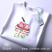 Футболка Їжте сало, біла, складена на поверхні, фон світлий, принт з милим шматком сала на дерев'яній дошці з ножем, текст під принтом яскравий і веселий.