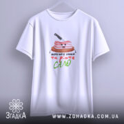 Футболка Їжте сало, біла, підвішена на вішалці, фон світло-сірий, принт з усміхненим шматком сала на дерев'яній дошці з ножем, текст під принтом кольоровий і привітний.