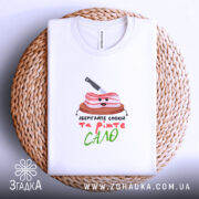 Футболка Їжте сало, біла, складена на кошику з ротанга, фон світлий, принт з веселим шматком сала на дерев'яній дошці з ножем, текст під принтом яскравий і привабливий.