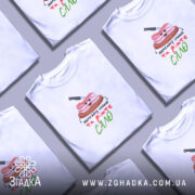 Футболка Їжте сало, біла, зображена на моделі, яка позує з легким усмішкою, фон м'який пастельний, принт з веселим шматком сала на дерев'яній дошці з ножем, текст під принтом написаний кольоровими літерами.