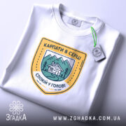 Футболка карпати в серці, біла, на столі, складена, з принтом у вигляді герба, що містить зображення гір, будинку та напис 'КАРПАТИ В СЕРЦІ СПОКІЙ У ГОЛОВІ', фон світлий, з елементами освітлення.