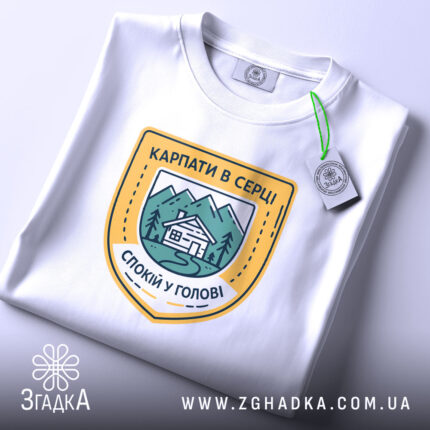 Футболка карпати в серці, біла, на столі, складена, з принтом у вигляді герба, що містить зображення гір, будинку та напис 'КАРПАТИ В СЕРЦІ СПОКІЙ У ГОЛОВІ', фон світлий, з елементами освітлення.