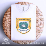 Футболка карпати в серці, біла, на плетеній кошику, з принтом у вигляді герба, що містить зображення гір, будинку та напис 'КАРПАТИ В СЕРЦІ СПОКІЙ У ГОЛОВІ', фон світлий, з текстурою кошика.