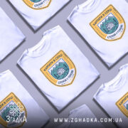 Футболка карпати в серці, біла, на моделі, з принтом у вигляді герба, що містить зображення гір, будинку та напис 'КАРПАТИ В СЕРЦІ СПОКІЙ У ГОЛОВІ', фон ніжно-фіолетовий.