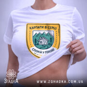 Футболка карпати в серці, біла, на столі, складена, з принтом у вигляді герба, що містить зображення гір, будинку та напис 'КАРПАТИ В СЕРЦІ СПОКІЙ У ГОЛОВІ', фон світлий, з елементами освітлення.