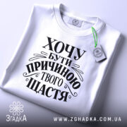 Футболка хочу бути причиною твого щастя, біла, розкладена на поверхні, з видимим принтом чорного кольору, текст оформлений в елегантному стилі, на фоні світло-сірого кольору, з зеленим ярликом.