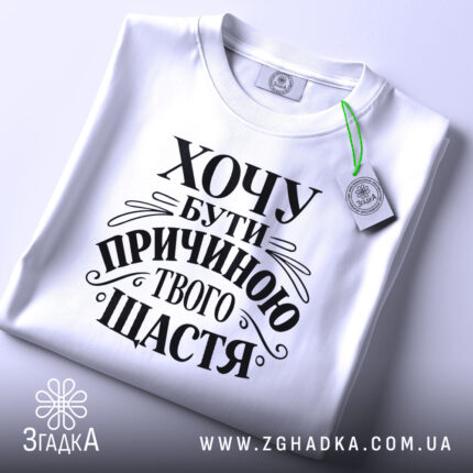 Футболка хочу бути причиною твого щастя, біла, розкладена на поверхні, з видимим принтом чорного кольору, текст оформлений в елегантному стилі, на фоні світло-сірого кольору, з зеленим ярликом.