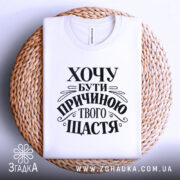 Футболка хочу бути причиною твого щастя, біла, складена на плетеній кошику, з чітким чорним принтом, текст оформлений в стильному шрифті, фон світлий, що створює затишну атмосферу.