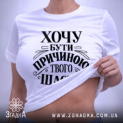 Футболка хочу бути причиною твого щастя, біла, розкладена на поверхні, з видимим принтом чорного кольору, текст оформлений в елегантному стилі, на фоні світло-сірого кольору, з зеленим ярликом.