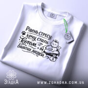 Футболка хочу стати котом. Біла футболка, зображена зверху, на плоскій поверхні. Принт чорного кольору з котом, який сидить на грошах, з текстом "Рівень стресу: хочу стати котом багатих людей". На футболці є етикетка з зеленим шнурком.