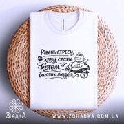 Футболка хочу стати котом. Біла футболка, зображена зверху на плетеному кошику. Принт чорного кольору з котом, який сидить на грошах, з текстом "Рівень стресу: хочу стати котом багатих людей".
