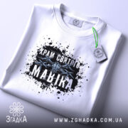 Футболка Храм святого Мавіка, біла, на столі, з принтом на грудях, фон світло-сірий, принт з чорними плямами та написом 'Храм святого Мавіка', етикетка з зеленим шнурком.