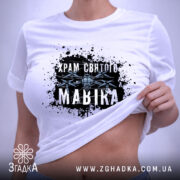 Футболка Храм святого Мавіка, біла, на столі, з принтом на грудях, фон світло-сірий, принт з чорними плямами та написом 'Храм святого Мавіка', етикетка з зеленим шнурком.