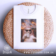 Футболка кіт спокій, біла футболка на плетеному кошику, з принтом кота в окулярах, що тримає чашку, на фоні з вогнем і вибухами, ракурс зверху, без моделі.