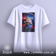 Футболка кіт за кермом, біла, з бокового ракурсу, підвішена на вішалці, фон з розмитими вогнями автомобільних фар, принт з котом за кермом з жовтою пов'язкою на голові, виразом радості на обличчі.