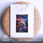 Футболка кіт за кермом, біла, з верхнього ракурсу, на плоскій поверхні, фон з плетеного кошика, принт з котом за кермом з жовтою пов'язкою на голові, виразом радості на обличчі.