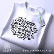 Футболка Кохання зле, біла, складена на поверхні, фон світло-сірий, принт чорного кольору з малюнками ангела та диявола, текстом "Кохання зле, полюбиш і мене", з етикеткою зеленого кольору.
