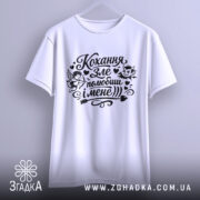 Футболка Кохання зле, біла, підвішена на вішалці, фон світло-сірий, принт чорного кольору з малюнками ангела та диявола, текстом "Кохання зле, полюбиш і мене".