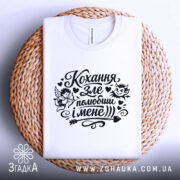 Футболка Кохання зле, біла, складена на плетеній основі, фон світло-сірий, принт чорного кольору з малюнками ангела та диявола, текстом "Кохання зле, полюбиш і мене".