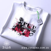 Футболка Королева випускного білого кольору, складена на поверхні, фон світлий, принт з чорними та червоними елементами, що зображає силует дівчини в короні, з етикеткою на зеленому шнурку.