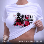 Футболка Королева випускного білого кольору, складена на поверхні, фон світлий, принт з чорними та червоними елементами, що зображає силует дівчини в короні, з етикеткою на зеленому шнурку.