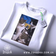 Футболка котяча банда, біла, з верхнього ракурсу, на столі, фон світло-сірий, принт з двома котами, один з яких тримає татуювальну машинку, на фоні жовто-блакитний прапор, з етикеткою на комірці.