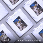 Футболка котяча банда, біла, з фронтального ракурсу, на моделі, фон світло-сірий, принт з двома котами, один з яких тримає татуювальну машинку, на фоні жовто-блакитний прапор.