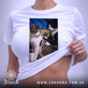 Футболка котяча банда, біла, з верхнього ракурсу, на столі, фон світло-сірий, принт з двома котами, один з яких тримає татуювальну машинку, на фоні жовто-блакитний прапор, з етикеткою на комірці.