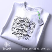 Футболка красива розумна, білого кольору, зображена на столі в горизонтальному положенні, фон світло-сірий, принт чорного кольору з написом "КРАСИВА РОЗУМНА, але кукуха підводить", на футболці є етикетка з логотипом.
