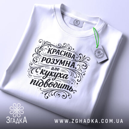 Футболка красива розумна, білого кольору, зображена на столі в горизонтальному положенні, фон світло-сірий, принт чорного кольору з написом "КРАСИВА РОЗУМНА, але кукуха підводить", на футболці є етикетка з логотипом.