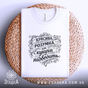 Футболка красива розумна, білого кольору, складена на плоскій поверхні, фон світло-сірий, принт чорного кольору з написом "КРАСИВА РОЗУМНА, але кукуха підводить", футболка розташована на плетеній основі.