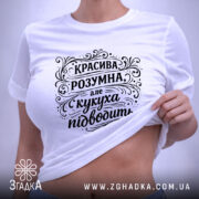 Футболка красива розумна, білого кольору, зображена на столі в горизонтальному положенні, фон світло-сірий, принт чорного кольору з написом "КРАСИВА РОЗУМНА, але кукуха підводить", на футболці є етикетка з логотипом.