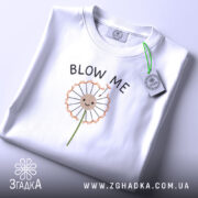 Футболка Кульбаба, біла, складена на поверхні, з принтом кульбаби та текстом 'BLOW ME', видно етикетку з зеленим шнурком, фон світлий.