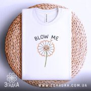 Футболка Кульбаба, біла, складена на плетеній основі, з принтом кульбаби та текстом 'BLOW ME', фон світлий.