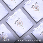 Футболка Кульбаба, біла, на моделі, з принтом у вигляді кульбаби з усміхненим обличчям, текстом 'BLOW ME' над принтом, фон ніжно-фіолетовий.