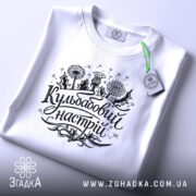 Футболка кульбабовий настрій, біла, складена на поверхні, ракурс зверху, фон світло-сірий, принт чорного кольору з написом 'Кульбабовий настрій' та малюнками квітів.