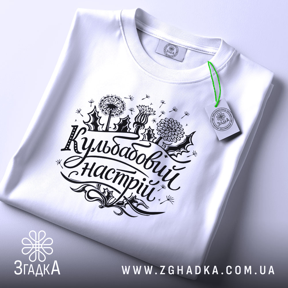 Футболка кульбабовий настрій, біла, складена на поверхні, ракурс зверху, фон світло-сірий, принт чорного кольору з написом 'Кульбабовий настрій' та малюнками квітів.