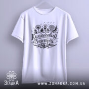 Футболка кульбабовий настрій, біла, підвішена на вішалці, ракурс спереду, фон світло-сірий, принт чорного кольору з написом 'Кульбабовий настрій' та ілюстраціями квітів.