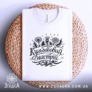 Футболка кульбабовий настрій, біла, складена на плетеній кошику, ракурс зверху, фон світло-сірий, принт чорного кольору з написом 'Кульбабовий настрій' та малюнками квітів.