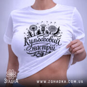 Футболка кульбабовий настрій, біла, складена на поверхні, ракурс зверху, фон світло-сірий, принт чорного кольору з написом 'Кульбабовий настрій' та малюнками квітів.