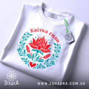 Футболка квітка сили, біла, складена на поверхні, фон світло-сірий, принт з червоним квіткою лотоса та зеленим листям, напис 'Квітка сили' вгорі, з етикеткою на шнурку.