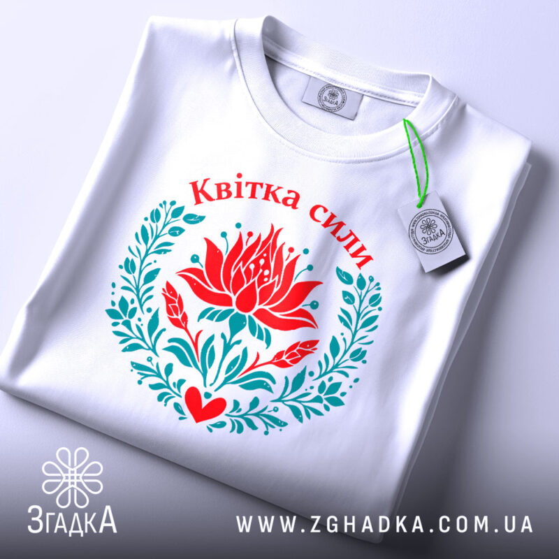 Футболка квітка сили, біла, складена на поверхні, фон світло-сірий, принт з червоним квіткою лотоса та зеленим листям, напис 'Квітка сили' вгорі, з етикеткою на шнурку.