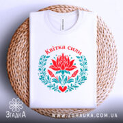 Футболка квітка сили, біла, складена на плетеній кошику, фон світло-сірий, принт з червоним квіткою лотоса та зеленим листям, напис 'Квітка сили' вгорі.