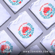 Футболка квітка сили, біла, зображена на моделі з бокового ракурсу, фон світло-сірий, принт з червоним квіткою лотоса та зеленим листям, напис 'Квітка сили' вгорі.