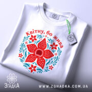 Футболка квітну бо жива, біла, лежить на поверхні, принт червоного квітки та зелених листків, фон світло-сірий, етикетка з зеленим шнурком.