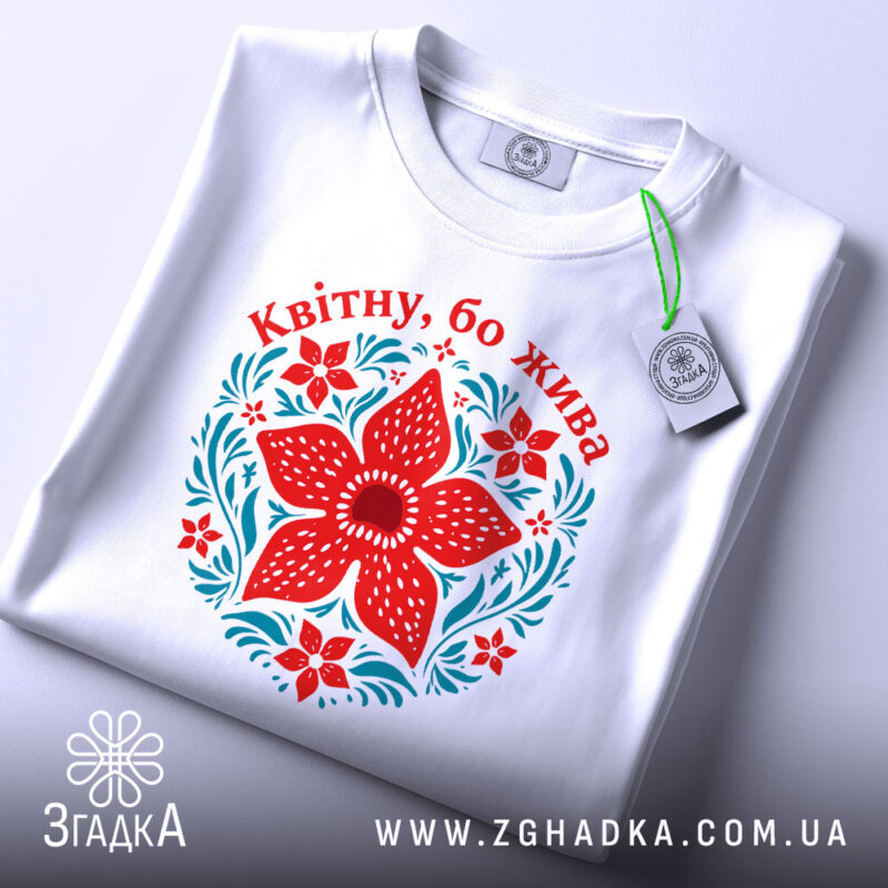 Футболка квітну бо жива, біла, лежить на поверхні, принт червоного квітки та зелених листків, фон світло-сірий, етикетка з зеленим шнурком.